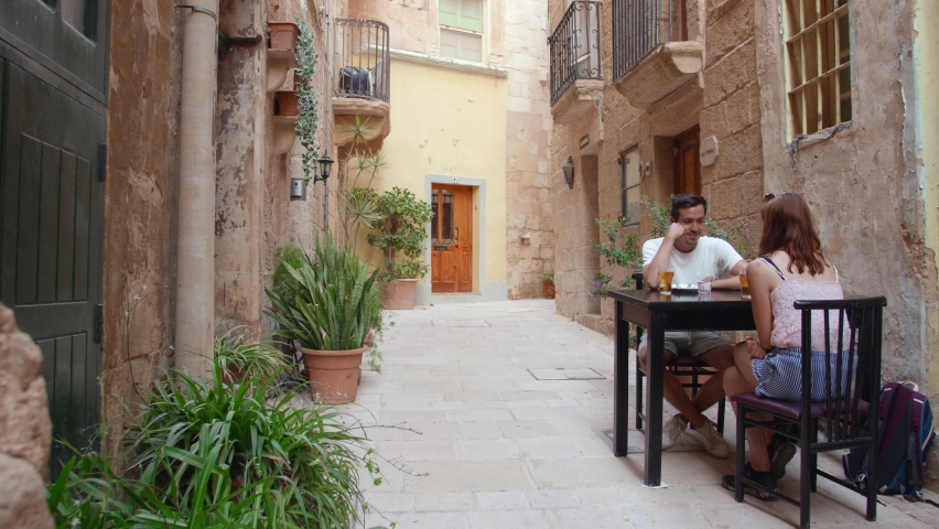 Couple Chilling And Drinking Beer On Table In Historic Street In Birgu, Malta.