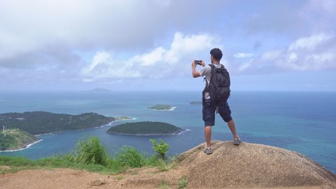 Young man with backpack stands on the high mountain at Phahindum view point popular landmark in Phuket Thailand and shoots video and photo with smartphone Concept vacation travel Phuket Thailand