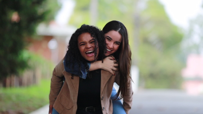 Two diverse girlfriends piggyback outside smiling and laughing