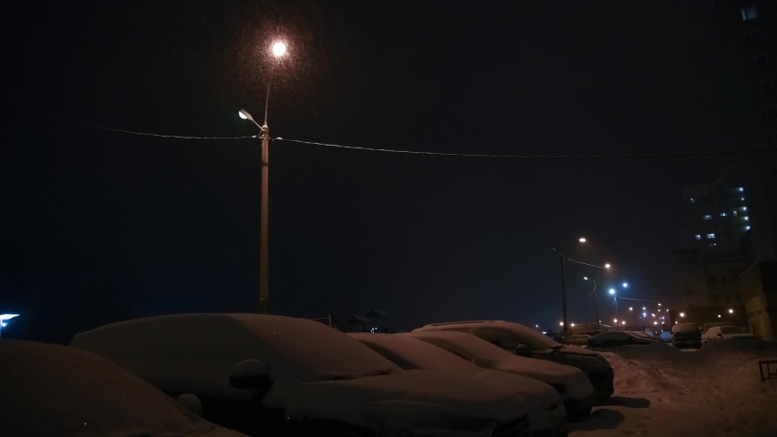 Selective focus. Heavy snow during blizzard with street lamp light at winter. Parked cars. Blurred apartment building in the background. 4K resolution video. Winter weather forecast theme.