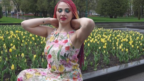 Awesome girl with rainbow braids and expressive glitter makeup. She sits and enjoys the warm spring weather in a green park with large tulip beds. UHD 4K 4x slow-motion video