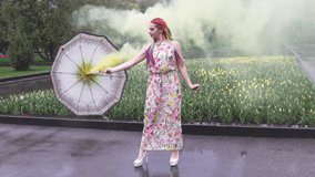 Adorable girl in a dress with a floral print with rainbow braids and makeup. Dances with an umbrella hiding in yellow smoke against the backdrop of flower bed with tulips. UHD 4K 4x slow-motion video