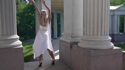   blonde woman is wearing gown of ancient greek goddess, posing near column of old palace, sexuality and femininity