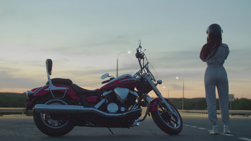 Woman stands next to her red cool motorcycle, removes the helmet from her head, holds it in her hands and looks at the track, back view