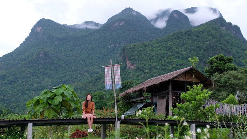 A young  female traveler sitting on wooden bridge with a beautiful mountains view on foggy day in background