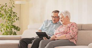 An aged couple sits on couch in living room, the husband sitting with glasses holds a tablet and taps his finger on it, the wife sitting next to him looks at the screen, they look at pictures, videos