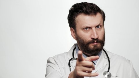 Doctor threatens with his finger. Serious frowning man doctor on white background looking at camera and points menacingly with his index finger