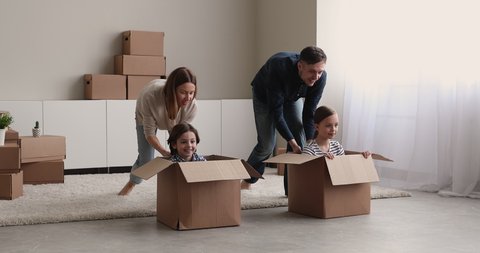 Happy young couple play with cute children at relocation day to new own dwelling, little kids sit inside carton boxes enjoy race playtime have fun at home. Overjoyed family celebrate move day concept