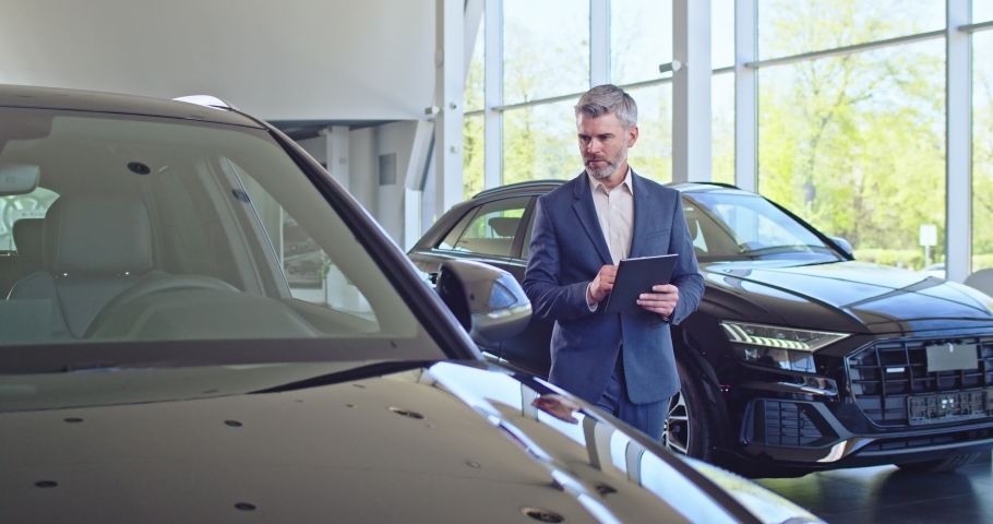 Serious caucasian salesman dressed in a business suit going between cars, looking around, using his tablet. Car dealership. Royalty-Free Stock Footage #1075331153