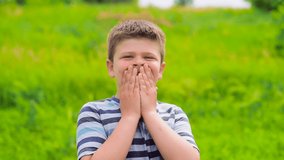 A beautiful boy of European appearance stands in a green field and laughs, covering his face with his hands. The child demonstrates sincere emotions of joy on his face. Slow-motion video, full hd