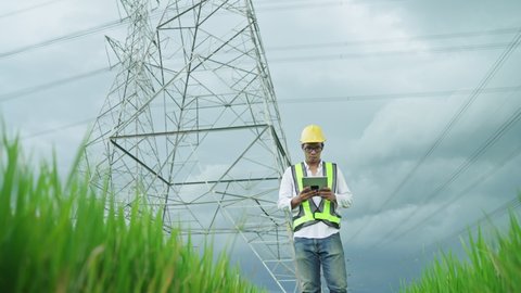 Font View.Electrical engineer wearing a Yellow helmet and safety carrying using tablet vest walking near high voltage electrical lines towards power station on the field.
