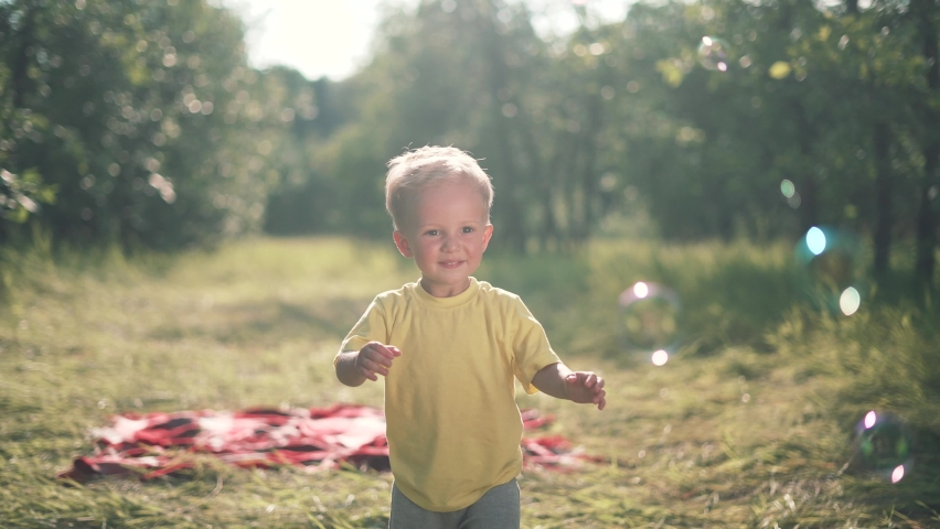 Happy kid is play in park. Baby play with soap bubbles. Kid smile and run through forest park.Smile of happy child. Baby smile and bursts soap bubbles. Child play.Baby run forest park for soap bubbles Royalty-Free Stock Footage #1075343084