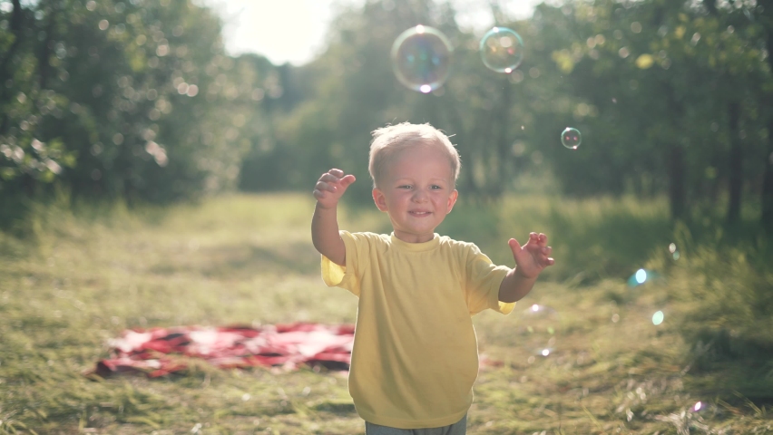 Happy kid is play in park. Baby play with soap bubbles. Kid smile and run through forest park.Smile of happy child. Baby smile and bursts soap bubbles. Child play.Baby run forest park for soap bubbles | Shutterstock HD Video #1075343084