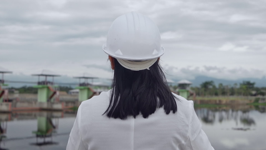 Female engineer in a white helmet reading data in a blueprint and checking the water system at the dam construction site to generate electricity.