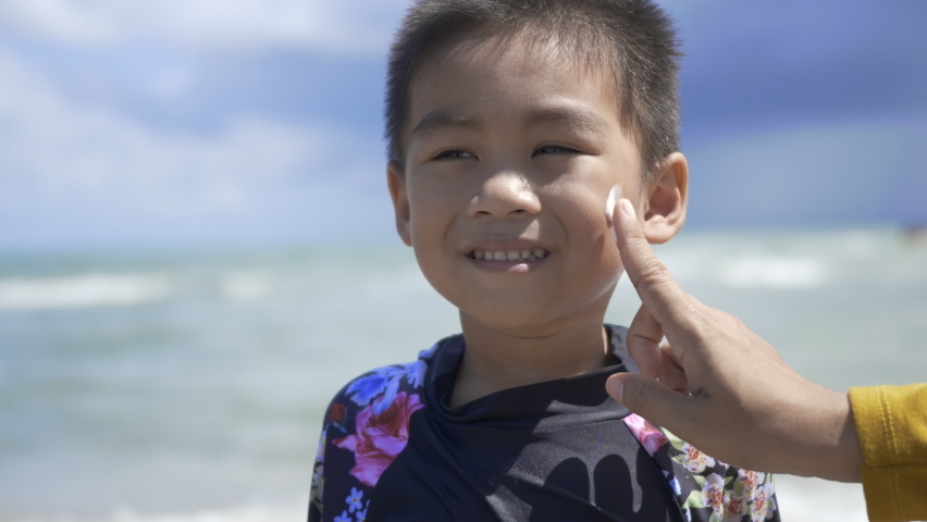 Happy little child boy standing at tropical beach during his summer vacation having sunscreen UV applied to face by mom, kid preschool before swimming activity summer vacation | Shutterstock HD Video #1075349303