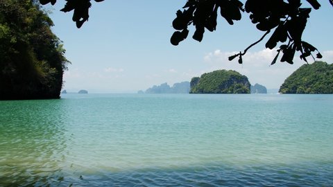 Limestone karsts dot the landscape of Phang Nga Bay, Phuket Thailand. The bright sun reflects on the blue turquoise water of the Andaman sea. Tree branches bounce in the soft tropical breeze.