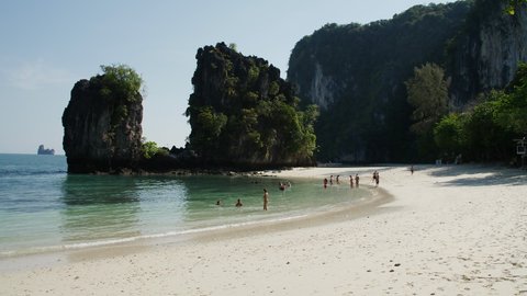 Tourists swim and play in the emerald green waves that gently lap onto the white sand beach of Hong Island, Krabi Thailand. Limestone karsts jut vertically out of the calm water in front of blue sky.
