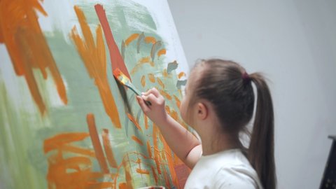 Kid girl with down syndrome draws with a brush on a large canvas in a white room, girl with special needs draws a color red-blue abstraction.