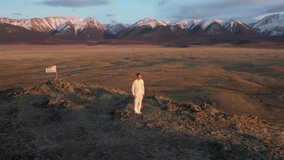 Video of kungfu master in a white sports uniform training wushu with jian sword on the hill. Man perform wushu forms in nature on background of snowy mountains at sunset time.