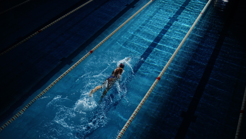 Female Swimmer Racing in Swimming Pool. Professional Athlete Overcoming Stress and Hardships in Dark Dramatic Pool, Cinematic Lap Lane Light Showing the Good Way. Aerial Slow Motion Shot Royalty-Free Stock Footage #1075356605