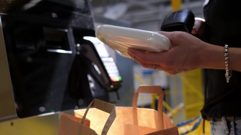 A woman browses through goods and puts purchases in a paper bag at a self-checkout counter. The buyer holds the scanner while scanning the item.