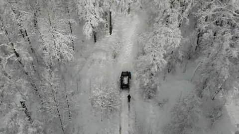 Aerial view of black SUV driving through deep white clean snow along trail in the mountains between trees. The rover gets stuck among snowy mountains and tall pines and firs. Extreme travel, tourism