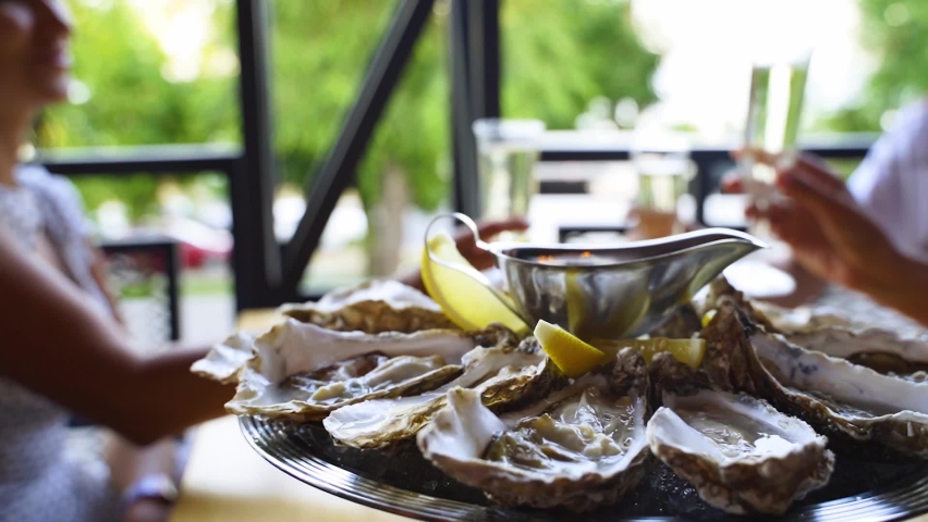 Luxury restaurant visitors clinking glasses with wine and cheering behind served plate of oysters on ice with lemon, sauce. Salver tray of traditional meditarian dish sea food for gourmets on a table. Royalty-Free Stock Footage #1075371728
