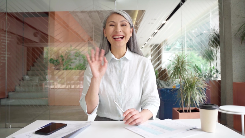 Smiling Asian middle aged businesswoman talking to camera at work by videocall conference, showing successful financial results, consulting client remotely online in modern office. Webcam view. Royalty-Free Stock Footage #1075373741