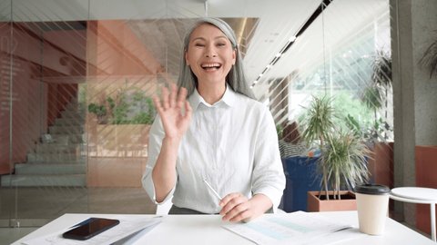 Smiling Asian middle aged businesswoman talking to camera at work by videocall conference, showing successful financial results, consulting client remotely online in modern office. Webcam view.