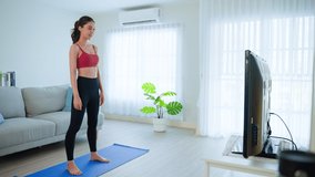 Asian Beautiful young woman stay home, doing aerobic exercise at home. Attractive girl doing lockdown activity, workout by squatting, follow instructions video from online trainer for health in house.