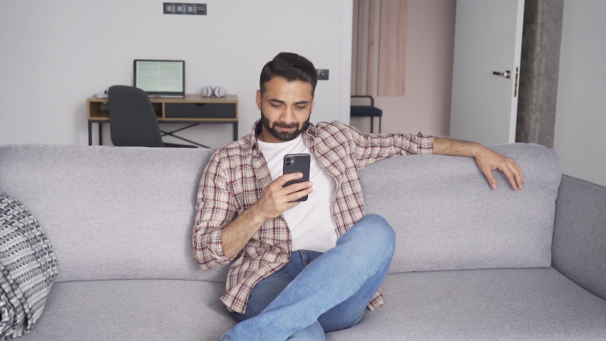 Young relaxed peaceful attractive indian eastern man sitting on coach in modern living room looking at cellphone checking social media, surfing internet, ordering delivery, reading ebook at weekend. | Shutterstock HD Video #1075375793