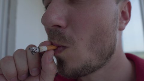 Young Man Smokes a Cigarette and Blows Out Smoke. Man's Lips Hold Cigarette with Orange Filter. Smoker and Harmful Lifestyle