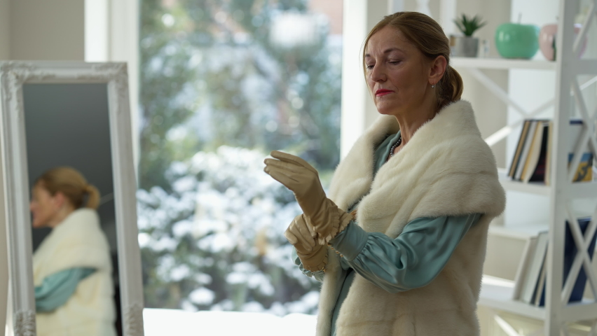 Medium shot portrait of confident elegant middle aged Caucasian lady putting on glove. Wealthy rich narcissistic woman getting ready for going out at home indoors. Style and fashion concept