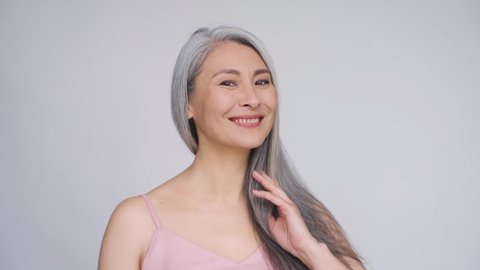 Gorgeous happy middle aged mature asian woman, senior older 50s lady smiling pampering touching hair waving grey healthy hair looking at camera. Ads of hair care, senior haircare advertising.
