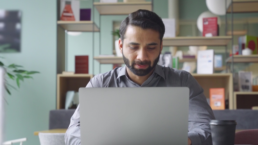 Young bearded indian businessman working on laptop in modern office lobby space. Indian executive using computer remote studying, watching online webinar, virtual training from homeoffice. Royalty-Free Stock Footage #1075378019
