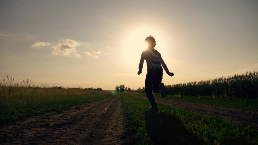 Happy kid run along rural road. Child run along road along wheat field. Happy child is playing at sunset. Silhouette of running kid on road at sunset. Kid running across the wheat field Royalty-Free Stock Footage #1075384292
