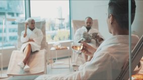 People relaxing at the spa salon. Video. Two men and a woman in white bathrobes having a rest with tea, newspaper inside a spacious room with panoramic windows.