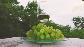 A hand pours white wine from a bottle into a glass that spins on a glass tray with a branch of green grapes. Creative slow motion video with speed up, down effect.	