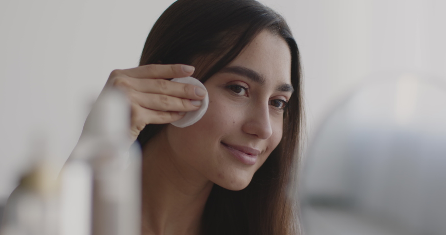 Make up removal and skin moisturizing. Young pretty eastern woman cleansing face with cotton pad, looking at mirror at bedroom and smiling, close up portrait, slow motion | Shutterstock HD Video #1075388843