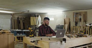 a European man with a beard and dark hair in a plaid shirt talking by video call on a laptop, explaining something, in a carpentry workshop, wooden products and tools visible