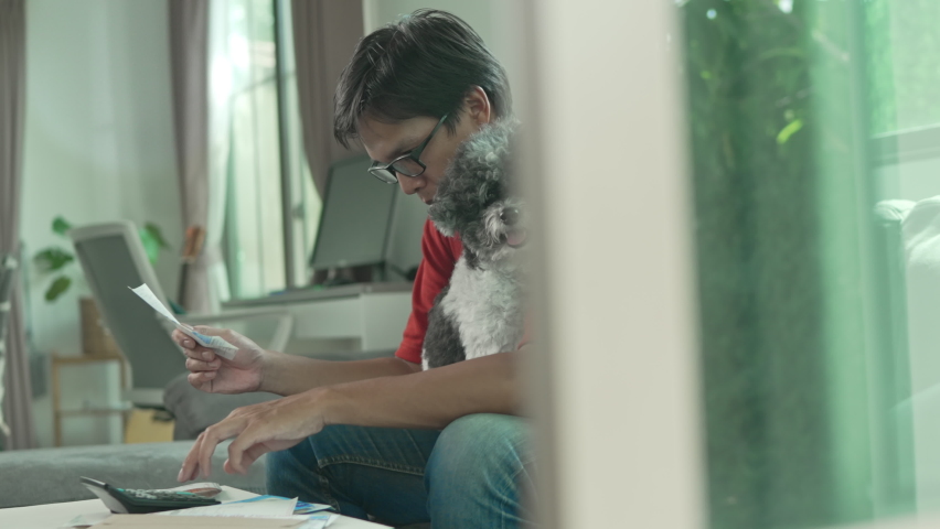 Asian man wear glasses red shirt and jeans with money and a calculator checks bills, calculates expenses, studies the credit balance sitting at the table with a dog at home,Concetp lifestlye finance.
 Royalty-Free Stock Footage #1075410638