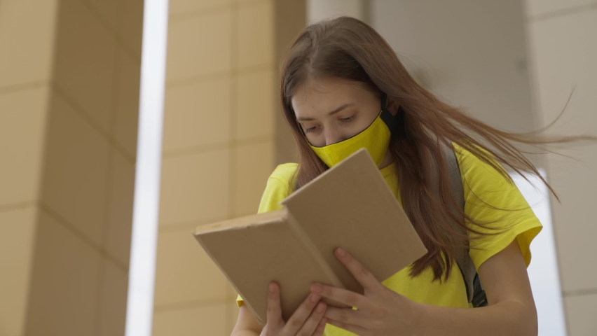 A schoolgirl girl in a mask reads a book while standing at the school, protection against coronavirus, covid-19 pandemic, air filtration through a mask filter, modern education for adolescents Royalty-Free Stock Footage #1075418858