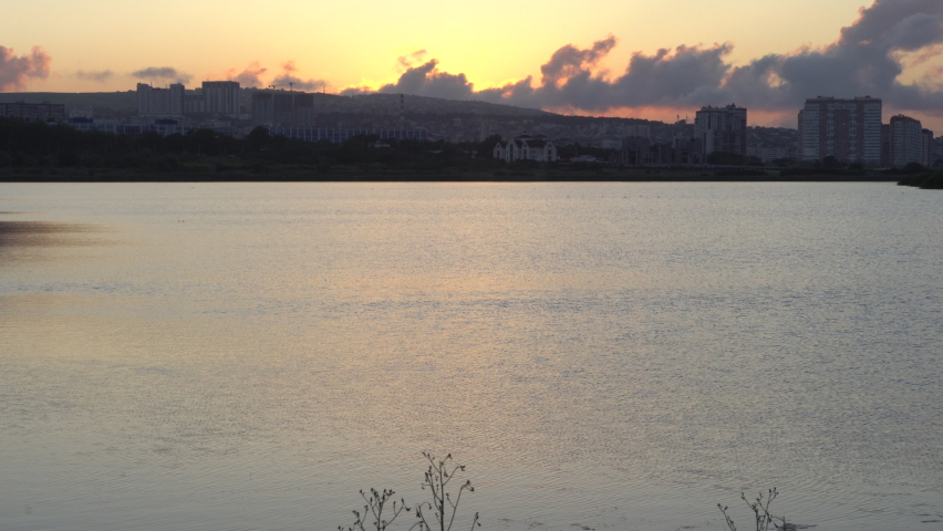 Big lake. Calm river. City landscape. Sun at sunset. Dark silhouettes. Dark clouds. Reflection in water. Golden sky. Ripples in the water. Panorama of the city.