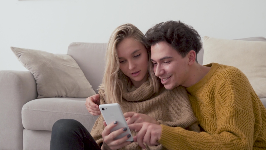 Young happy couple having fun using smartphone at home. Girl and guy talking, laughing, looking at mobile phone buying retail products shopping online, ordering delivery, checking social media apps. Royalty-Free Stock Footage #1075423748
