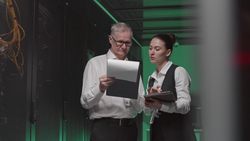 Medium long of senior Caucasian male and young female IT-specialists wearing white shirts and suits, standing in server room, holding documents and tablet computer, talking