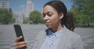 Distant communication. Close up portrait of cheerful african american business lady video chatting with partners via smartphone, standing outdoors in urban park, tracking shot