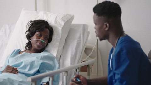 An African male doctor interviews a patient lying in a hospital bed with an oxygen mask. A black woman lying in a hospital bed describes the symptoms to the doctor