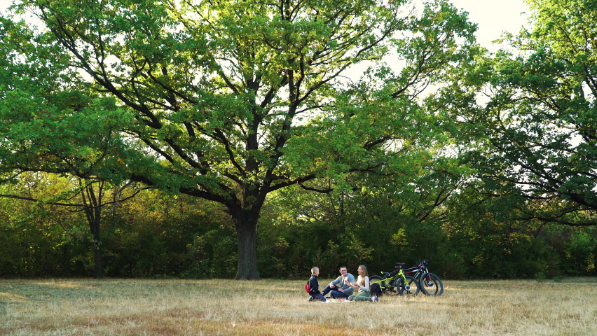 Happy family sitting on ground under big oak tree and clapping hands together, bicycles standing nearby. Parents and child relaxing after bike ride in nature. Concept of picnic