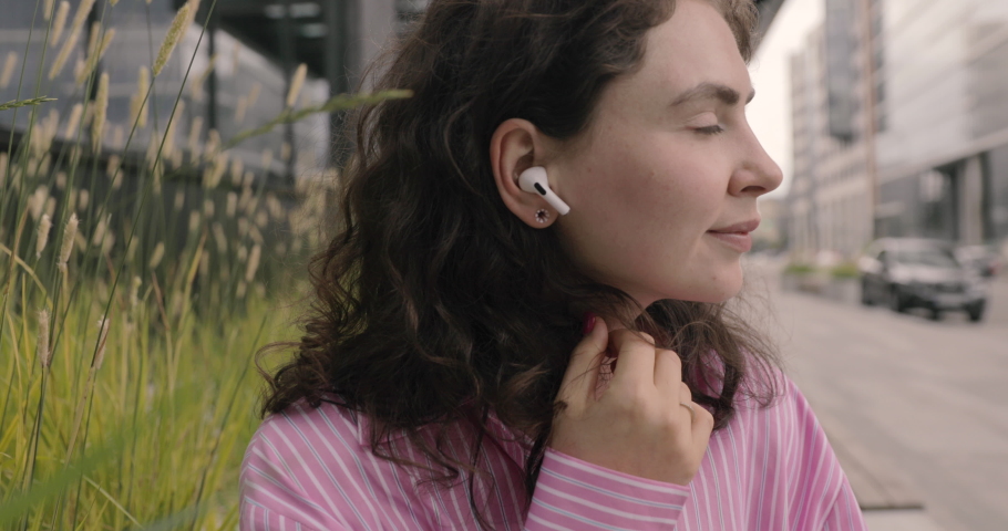 Attractive young woman listening the music in wireless in-ear headphones use smartphone. Apple AirPods Pro. Walk in the city, sit and look around. Smile portrait.