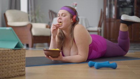 Careless obese woman lying on exercise mat eating chips listening to music in headphones dancing. Portrait of unmotivated Caucasian overweight lady enjoying hobby instead of training